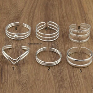 Puede incluir: Seis anillos de plata para dedos de los pies se muestran sobre una superficie de madera. Los anillos tienen varios diseños, incluyendo múltiples bandas, una forma de V y una pequeña bola. Cada anillo está numerado para su identificación.