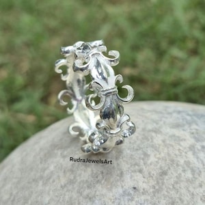Può includere: Un anello d'argento con un design a giglio. L'anello presenta un motivo ripetuto di emblemi floreali stilizzati. L'anello è su una superficie di pietra grigia. Il testo "RudraJewelsArt" è visibile.