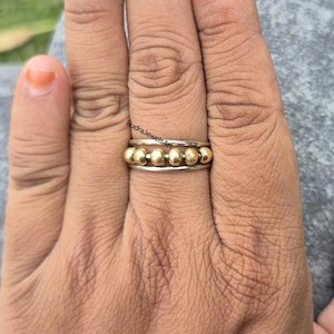 Peut inclure: Une bague en argent ornée de six perles dorées. La bague a un design simple avec une bande argentée et les perles dorées sont espacées uniformément sur le dessus. La bague est sur le doigt d'une personne.