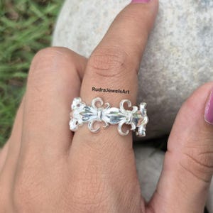 May include: A silver ring featuring a repeating fleur-de-lis design. The ring is worn on a finger, showcasing the intricate details of the floral pattern. The ring's surface reflects light, highlighting its polished finish. The text "RudraJewelsArt" is visible.