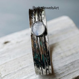 Könnte beinhalten: Ein silberfarbener Spinner-Ring mit einem eingravierten Blumenmuster und einem zentralen, runden, irisierenden Mondstein. Der Ring hat ein strukturiertes Design und der Text "RudrajewelsArt" ist sichtbar.