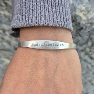Może przedstawiać: Srebrna bransoletka z grawerowanym napisem "RudraJewelsArt" z przodu. Bransoletka jest noszona na nadgarstku, z szarym rękawem swetra widocznym powyżej.