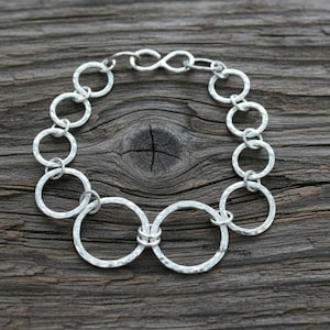 May include: A silver bracelet with a hammered texture, featuring a series of interconnected circles and chain links. The bracelet is displayed on a rustic wooden surface, showcasing its handcrafted design and metallic shine.