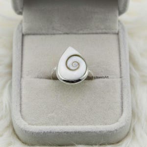 Op de afbeelding: Een zilveren ring met een druppelvormige, witte edelsteen met een spiraalpatroon. De ring is gezet in een zilveren band en wordt gepresenteerd in een grijze sieradendoos.