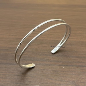 May include: A silver-colored double-band cuff bracelet. The bracelet has a smooth, polished finish and a minimalist design. The open-ended design allows for easy wear. The bracelet is displayed on a brown wooden surface. The text "RudraJewelsArt" is visible.