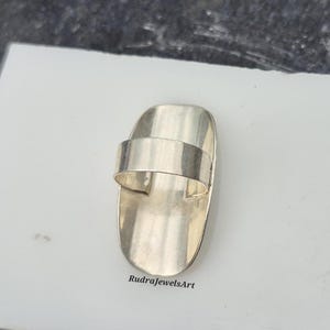 May include: A silver-toned ring with a rectangular band and a wide, curved top. The ring has a smooth, polished finish and is set against a white surface. The text "RudraJewelsArt" is visible on the surface.