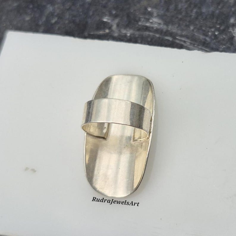 May include: A silver-toned ring with a rectangular band and a wide, curved top. The ring has a smooth, polished finish and is set against a white surface. The text "RudraJewelsArt" is visible on the surface.