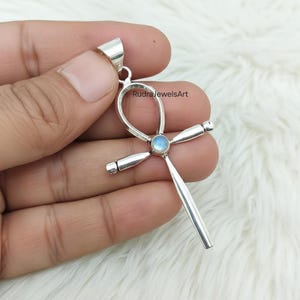 Könnte beinhalten: Ein silberner Ankh-Anhänger mit einem kleinen, runden, hellblauen Edelstein in der Mitte. Der Anhänger wird in einer Hand vor einem weißen, flauschigen Hintergrund gehalten. Der Text "RudraJewelsArt" ist sichtbar.