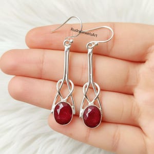 Puede incluir: Un par de pendientes de plata con un diseño largo y delgado. Cada pendiente presenta una piedra preciosa ovalada de color rojo intenso. Los pendientes cuelgan de un gancho simple, sobre un fondo blanco.