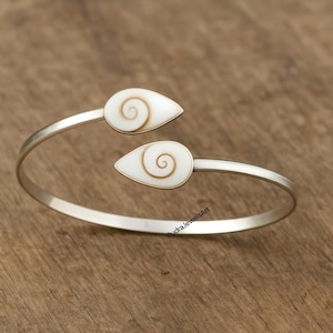 Może przedstawiać: Srebrna bransoletka na nadgarstek z dwoma białymi wstawkami w kształcie łezki, każda z spiralnym wzorem w ciepłym odcieniu. Otwarta bransoletka jest zaprojektowana z myślą o łatwym noszeniu. Prosty i elegancki design.