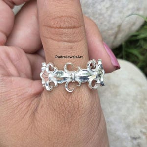 May include: A silver ring with a fleur-de-lis design. The ring features a repeating pattern of stylized floral elements. The ring is worn on a finger. The text "RudraJewelsArt" is visible.