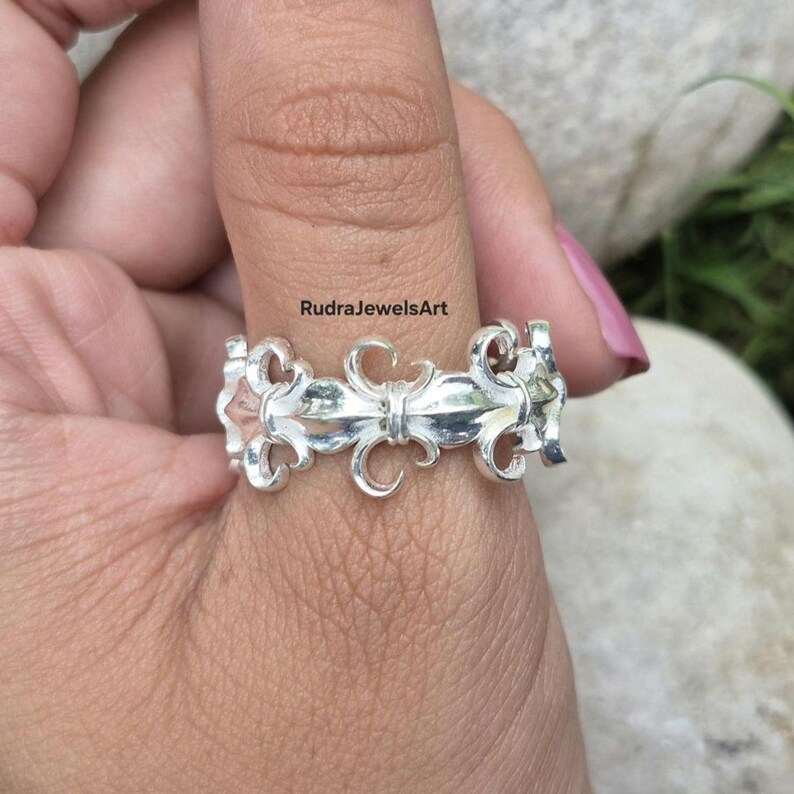 May include: A silver ring with a fleur-de-lis design. The ring features a repeating pattern of stylized floral elements. The ring is worn on a finger. The text "RudraJewelsArt" is visible.
