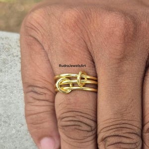May include: A gold-colored ring with a knot design. The ring has three bands that are intertwined to create the knot. The ring is on a finger. The text "RudraJewelsArt" is in the upper left corner.