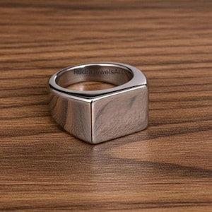May include: A silver-coloured, square-faced ring with a polished finish. The ring has a flat top and a thick band. The text "RudraJewelsArt" is visible on the ring. The ring is displayed on a wooden surface.
