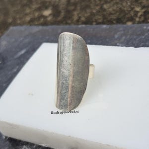 May include: A silver-toned ring with a long, rectangular face. The face of the ring has a textured, brushed metal appearance. The ring is set on a white surface. The text "RudraJewelsArt" is visible.