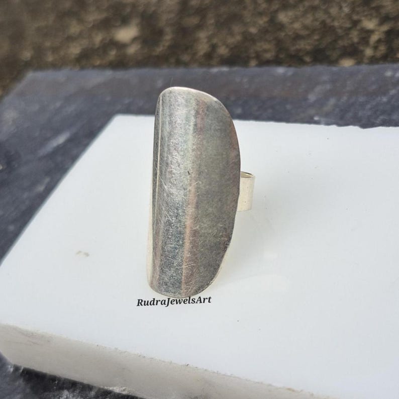 May include: A silver-toned ring with a long, rectangular face. The face of the ring has a textured, brushed metal appearance. The ring is set on a white surface. The text "RudraJewelsArt" is visible.