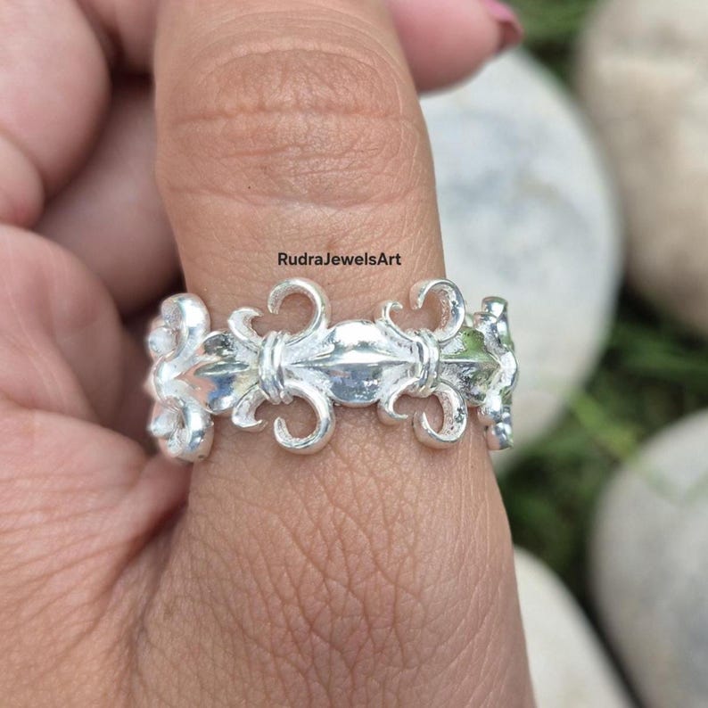 May include: A close-up of a silver ring with a repeating fleur-de-lis design. The ring is worn on a finger and has a polished finish. The ring's design is symmetrical and detailed, with curved elements.