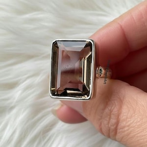 May include: A rectangular smoky quartz gemstone ring with a silver band. The gemstone is a light brown color and is set in a rectangular silver bezel. The ring is held up by a person's fingers.