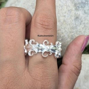 May include: A silver ring with a fleur-de-lis design. The ring features a repeating pattern of stylized floral motifs. The ring is worn on a finger. The text "RudraJewelsArt" is visible in the background.