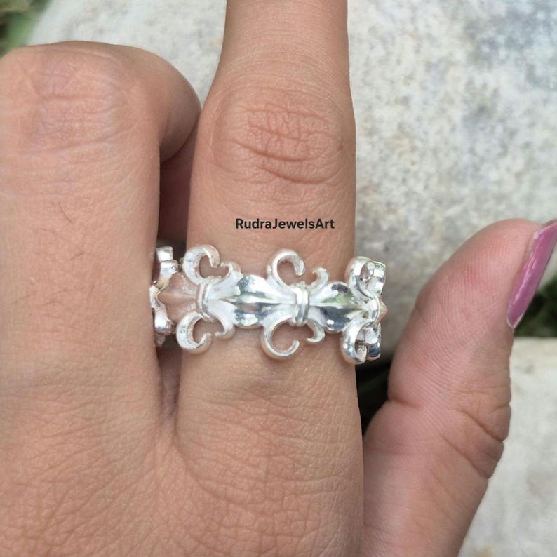 May include: A silver ring with a fleur-de-lis design. The ring features a repeating pattern of stylized floral motifs. The ring is worn on a finger. The text "RudraJewelsArt" is visible in the background.