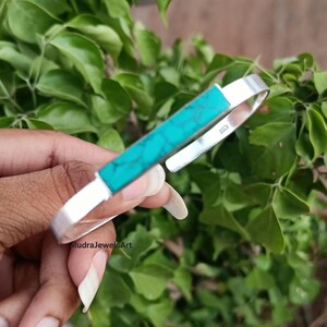 May include: A silver cuff bracelet with a rectangular turquoise stone inlay. The bracelet is held against a backdrop of green foliage. The text "RudraJewelsArt" is visible.