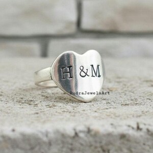 Può includere: Anello d'argento con una faccia a forma di cuore. Le lettere "H & M" sono stampate sul cuore. L'anello è su una superficie grigio chiaro. Il testo "RudraJewelsArt" è nella parte inferiore del cuore.