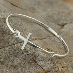 Könnte beinhalten: Ein silberfarbenes Armreif-Armband mit Kreuz-Design. Das Armband hat ein schlichtes, elegantes Design mit einem Kreuz als Mittelpunkt. Der Text "RudraJewelsArt" ist auf dem Armband zu sehen.