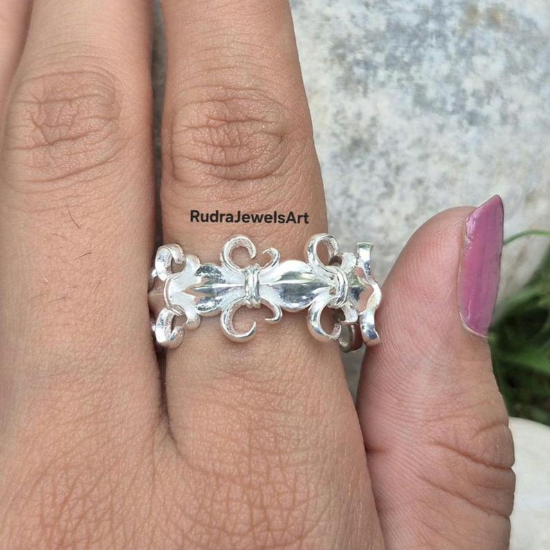 May include: A silver ring featuring a fleur-de-lis design. The ring is held by a hand with a pink painted fingernail. The text "RudraJewelsArt" is visible in the image.