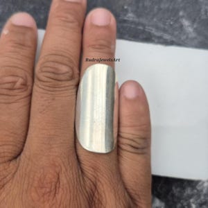 May include: A silver-colored ring with a rectangular design is worn on a finger. The ring has a smooth, polished surface. The text "RudraJewelsArt" is visible on the image.