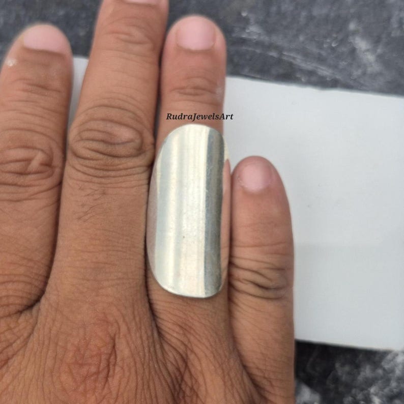 May include: A silver-colored ring with a rectangular design is worn on a finger. The ring has a smooth, polished surface. The text "RudraJewelsArt" is visible on the image.