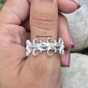 May include: A silver ring featuring a repeating fleur-de-lis design. The ring is worn on a finger, showcasing the intricate details of the floral pattern. The ring's surface reflects light, highlighting its polished finish. The text "RudraJewelsArt" is visible.