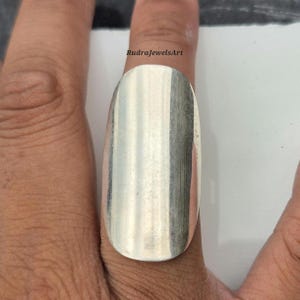 May include: A large, silver-coloured ring with an elongated oval shape. The ring is worn on a finger, and the surface reflects light, creating a striped pattern. The text "RudraJewelsArt" is visible.