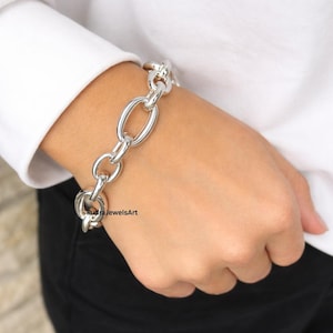 Könnte beinhalten: Ein silberfarbenes Kettenarmband mit ovalen und runden Gliedern. Das Armband wird an einem Handgelenk getragen, mit einem weißen Langarmhemd im Hintergrund. Das Armband hat einen klobigen Stil.
