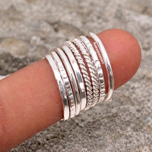 May include: A collection of silver rings of varying designs, including smooth bands, textured bands, and twisted rope-like rings. The rings are stacked on a finger, showcasing the different styles and textures. The brand name "RudrajewelsArt" is visible on one of the rings.