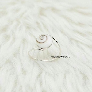 Può includere: Un anello d'argento con una pietra a forma di spirale bianca e marrone. La fascia dell'anello si curva attorno al dito, con la pietra posta in cima. L'anello è esposto su una superficie morbida, bianca e strutturata.