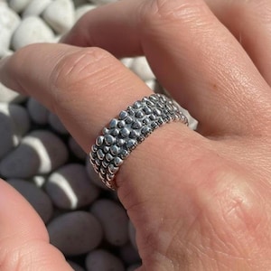 May include: A silver ring with a textured surface, featuring a pattern of small, raised, rounded shapes. The ring is worn on a finger, showcasing its design and craftsmanship. The ring is a piece of jewelry.