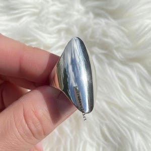 May include: A polished silver ring with a smooth, elongated design. The ring has a rounded top and tapers to a point. The text "BudraJewelsArt" is visible on the side of the ring.