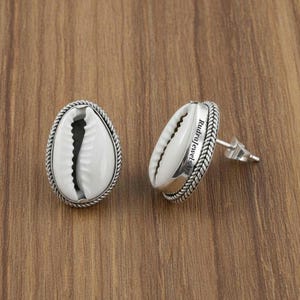 Puede incluir: Pendientes de plata con forma de concha de cauri blanca. Las conchas están engastadas en un marco ovalado de plata con un diseño de cuerda trenzada. Los pendientes tienen cierre de tuerca.