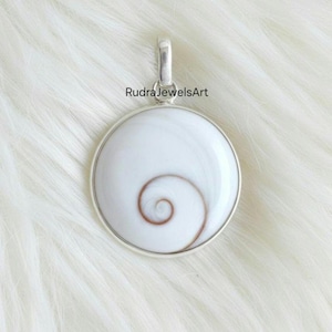 May include: A round, white pendant with a spiral design in the center, set in a silver-colored metal frame. The pendant has a loop at the top for a chain. The text "RudraJewelsArt" is visible.