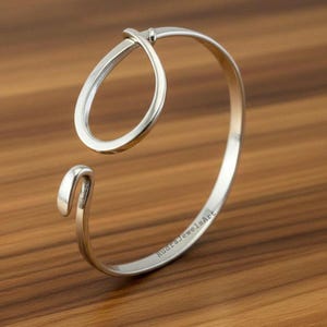 May include: A silver-colored open cuff bracelet with a teardrop-shaped design. The bracelet has a smooth, polished finish and the text "RudraJewelsArt" is engraved on the inside. The bracelet is displayed on a wooden surface.