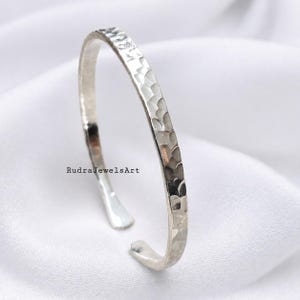 Puede incluir: Una pulsera de plata con textura martillada. La pulsera abierta se muestra sobre una superficie blanca. El texto "RudraJewelsArt" es visible en la superficie.