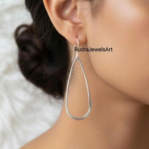 May include: Silver teardrop earrings. The earrings are a simple, elegant design with a smooth, polished finish. The earrings are lightweight and dangle from a hook. The brand name "RudraJewelsArt" is visible.