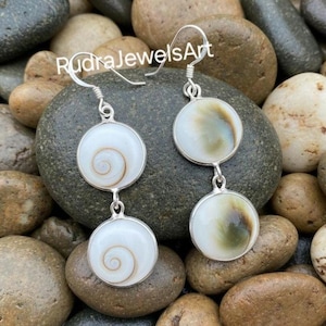 May include: Silver dangle earrings with two circular pendants. The top pendant is a light green and white color, while the bottom pendant is white with a brown spiral design. The earrings are displayed on a bed of rocks.