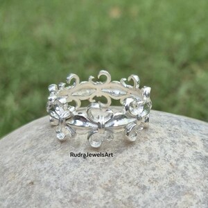 May include: A silver ring with an openwork design featuring floral and scroll motifs. The ring is set on a stone surface with a green background. The text "RudraJewelsArt" is visible on the stone.