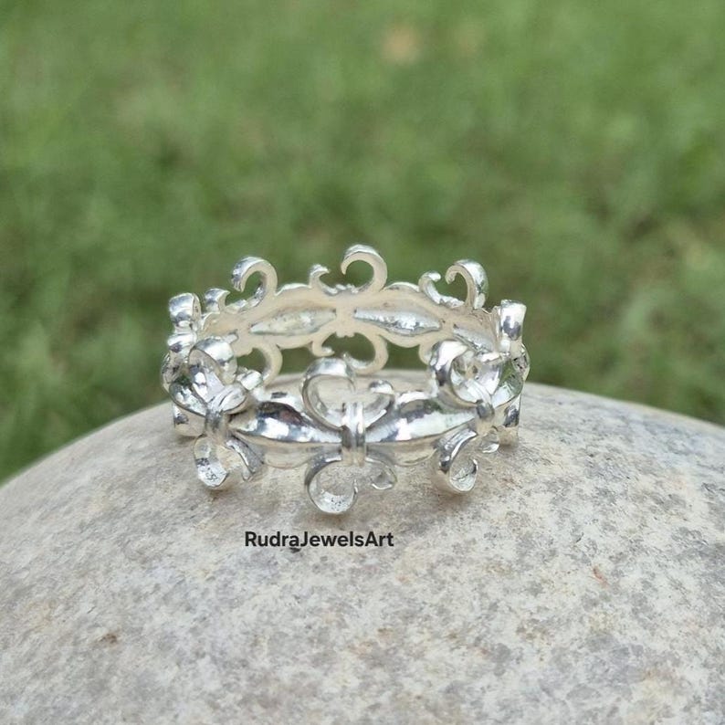May include: A silver ring with an openwork design featuring floral and scroll motifs. The ring is set on a stone surface with a green background. The text "RudraJewelsArt" is visible on the stone.