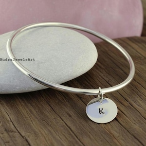 Puede incluir: Una pulsera de plata con un dije circular. El dije tiene la letra "K". La pulsera se muestra sobre una superficie de madera, con una piedra gris y una piedra rosa en el fondo. El texto "RudraJewelsArt" es visible.