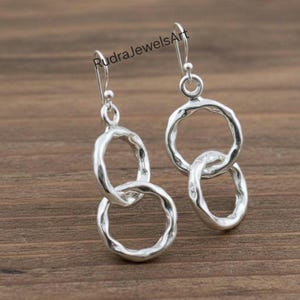 Puede incluir: Pendientes colgantes plateados con un diseño de doble círculo enlazado. Los pendientes tienen un aspecto texturizado y martillado y están suspendidos de un gancho simple. Los pendientes se exhiben sobre una superficie de madera.