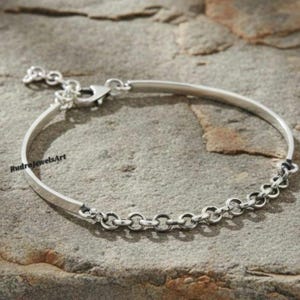 May include: A silver bracelet with a bar and chain design. The bracelet has a curved bar with a clasp and a chain of small, connected circles. The bracelet is displayed on a textured, gray surface.