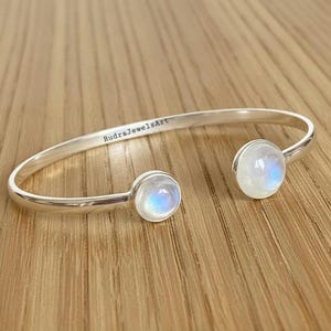 May include: A silver open cuff bracelet featuring two round moonstone cabochons. The bracelet is engraved with "RudraJewelsArt". The moonstones have a blue and white iridescence. The bracelet is displayed on a wooden surface.