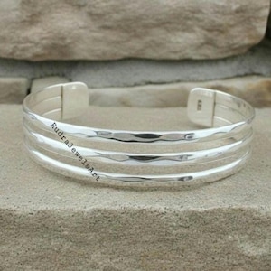 May include: A silver cuff bracelet with three textured bands. The bracelet has a hammered finish and the text "RudraJewelsArt" is visible on the side. The bracelet is displayed on a stone surface.
